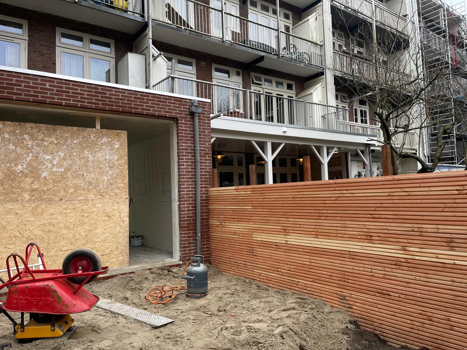 Vooraanzicht: de gevel staat, pui-openingen dichtgezet met OSB, terras nog een zandbed.