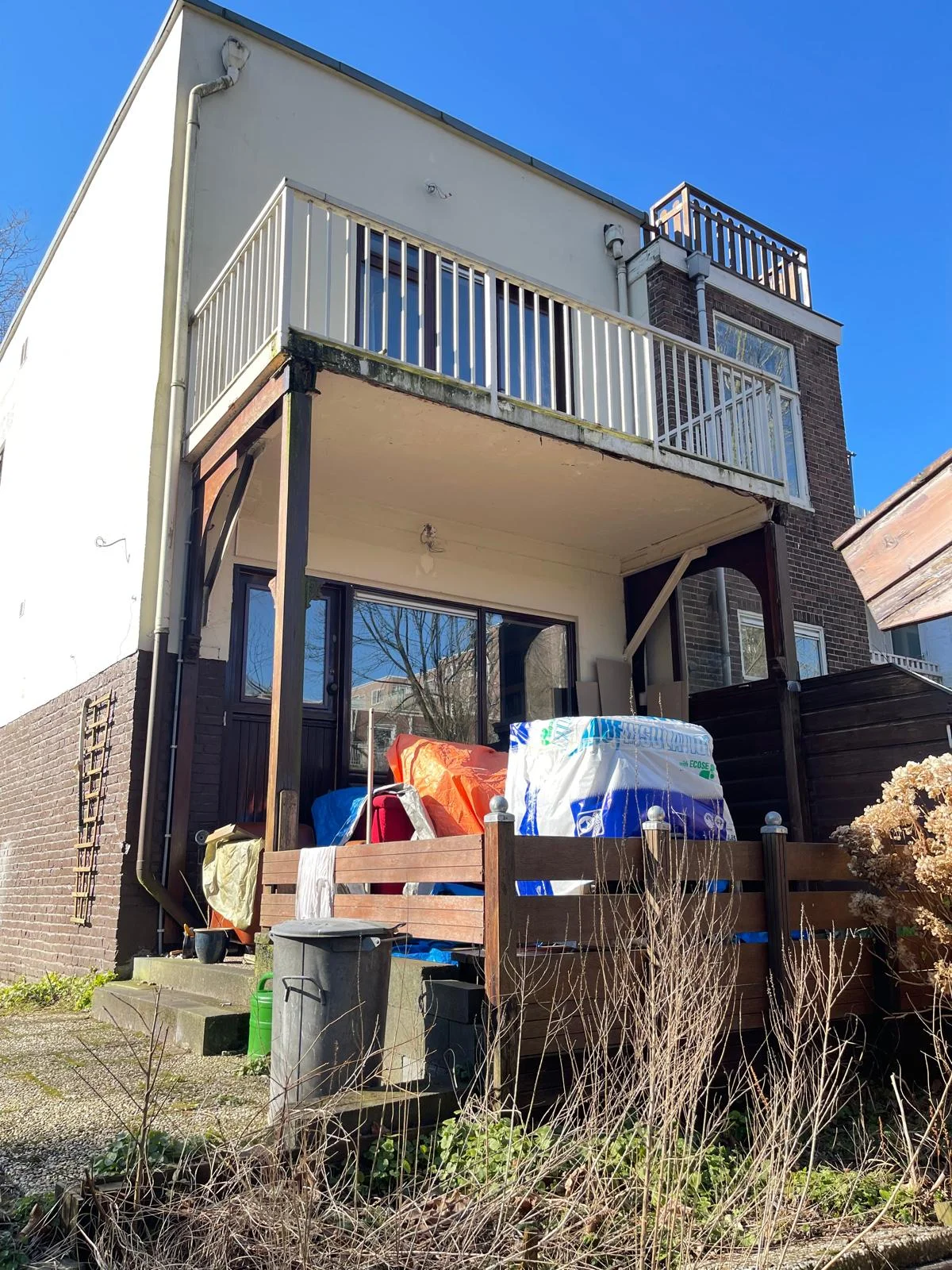 De achterzijde van de woning vóór de aanbouw: uitkragend balkon op de eerste verdieping, één tuindeur op de begane grond en opslag van tuinmeubels onder het balkon.