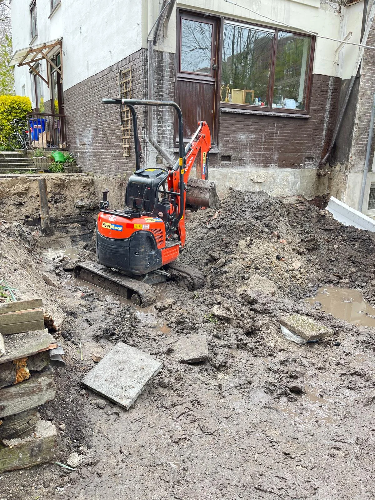 De minigraver opent het talud naast de bestaande gevel; sleuven die de toekomstige funderingsstroken volgen.