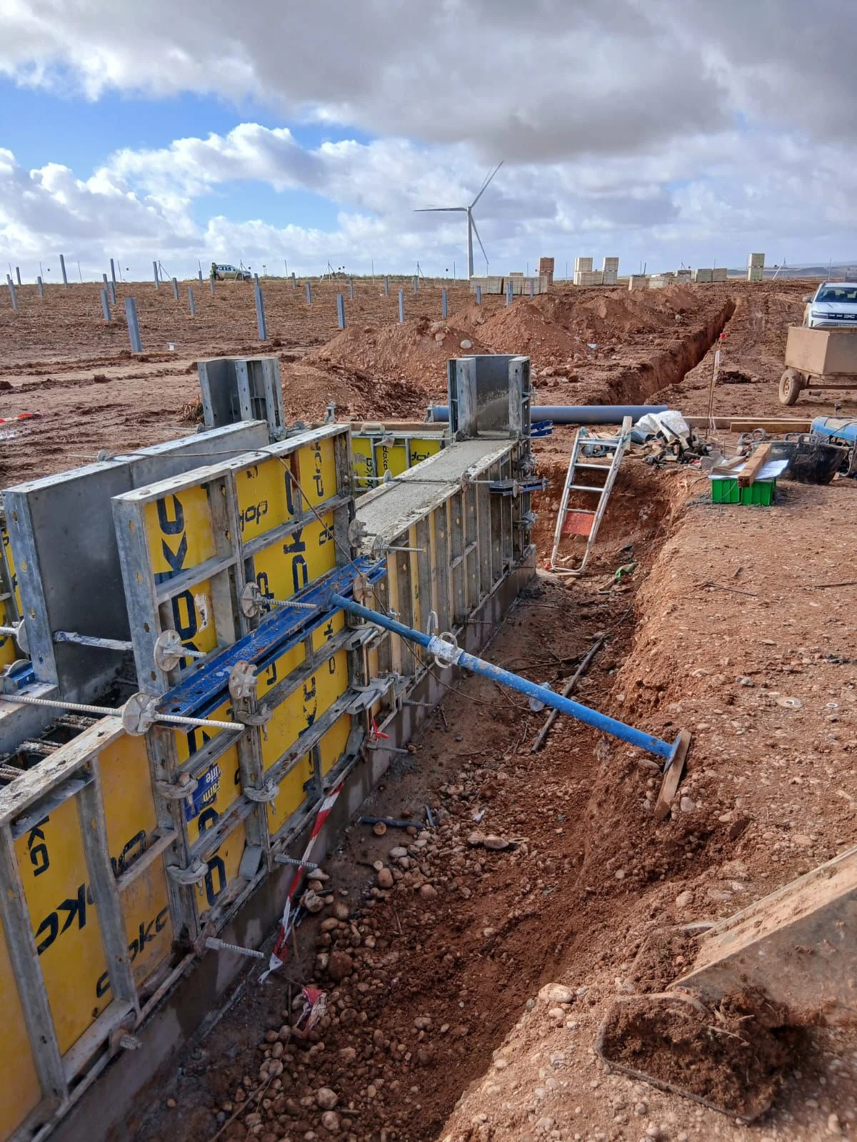 Geel-metalen modulair bekistingssysteem met rode diagonale stempels, klaar voor een betonstort; één windturbine op de achtergrond.