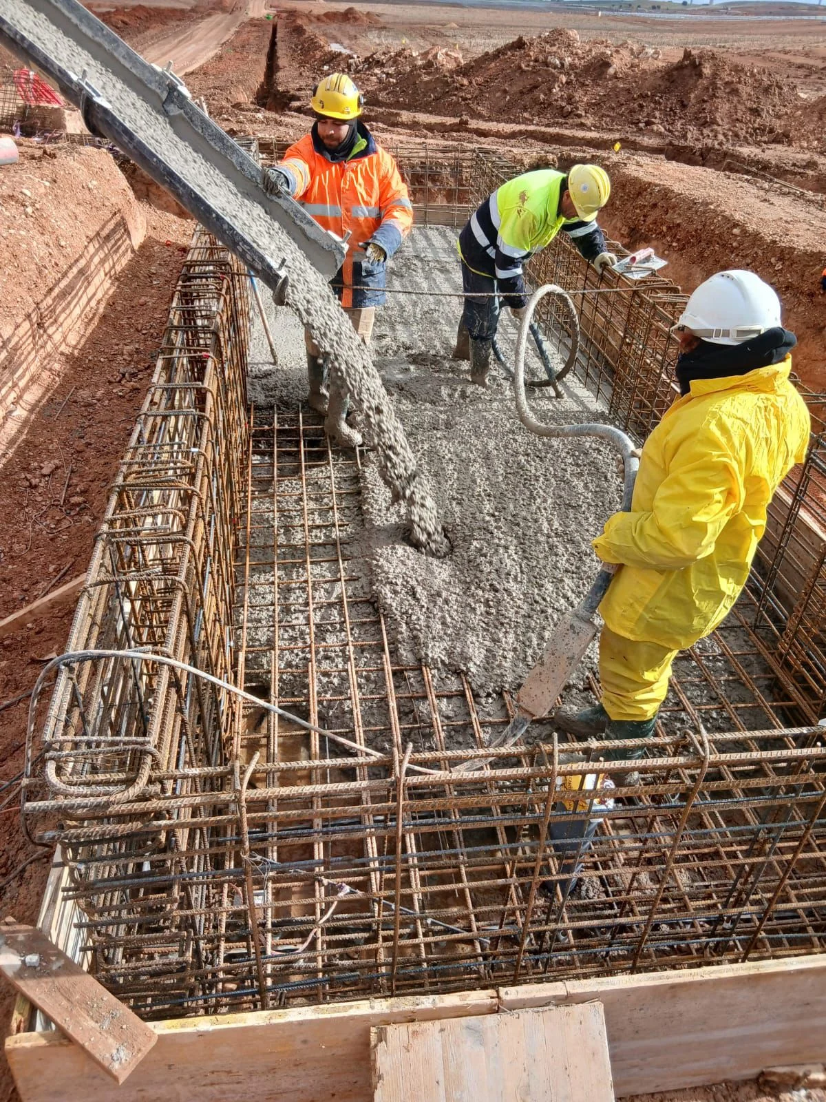 Betonpomp-arm stort vloeibare beton in de wapeningskorf; drie vakmannen in volledige PBM controleren het stortpatroon.
